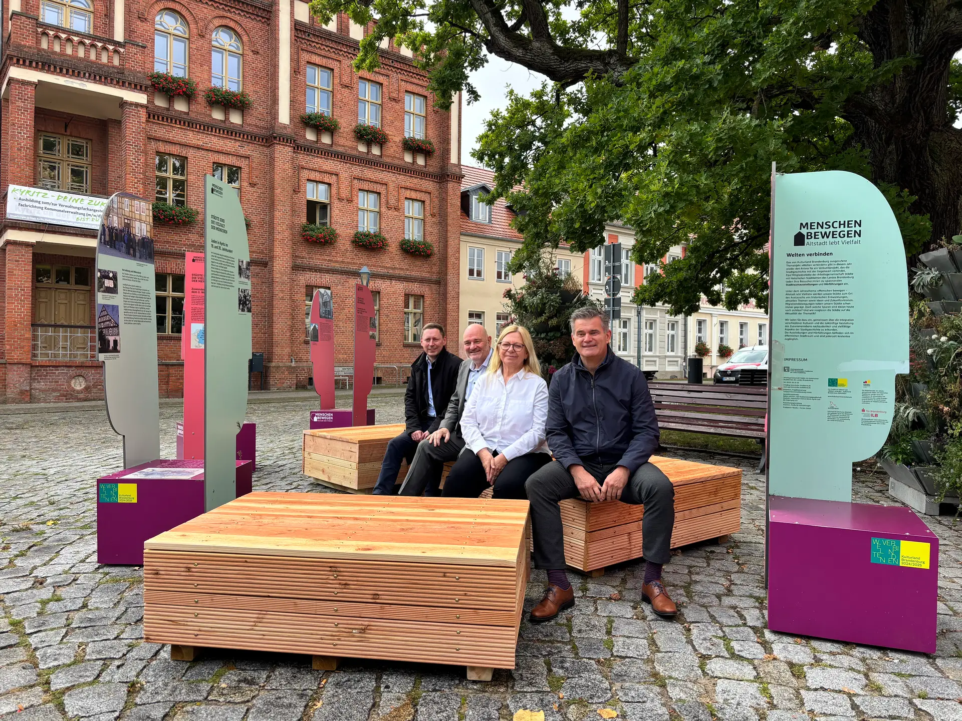 Auf einem Foto sitzen mehrere Personen auf neuen Stadtmöbeln; im Hintergrund ist eine Veranstaltungssituation im Rahmen eines Jahresforums zu erkennen.
